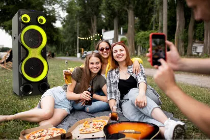 3 amigas sentadas na grama, tirando foto, comendo pizza, sorrindo e curtindo, com várias árvores de fundo e uma caixa de som torre da Polyvox ao lado delas