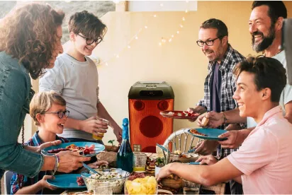 Família com 6 integrantes almoçando e se divertindo juntos, em uma mesa com bastante comida e com uma caixa de som Polyvox tocando música