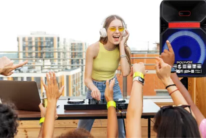 DJ mulher branca de cabelo liso, controlando uma mesa de som com uma caixa de som polyvox ao lado dela em um pedestal, e várias pessoas com a mão pro alto curtindo a música