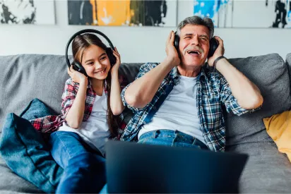 Avô e neta sentados em um sofá na sala, assistindo um vídeo no notebook, com fones de ouvido headset da Polyvox na cabeça, rindo e achando graça do que estão escutando