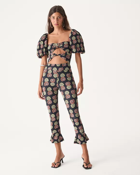 A Modelo esta vestindo uma Calça corsário feminina com estampa de conchas, cós alto e babado nas barras, inspirada no estilo retrô dos anos 40 e 50, ideal para looks sofisticados de verão.